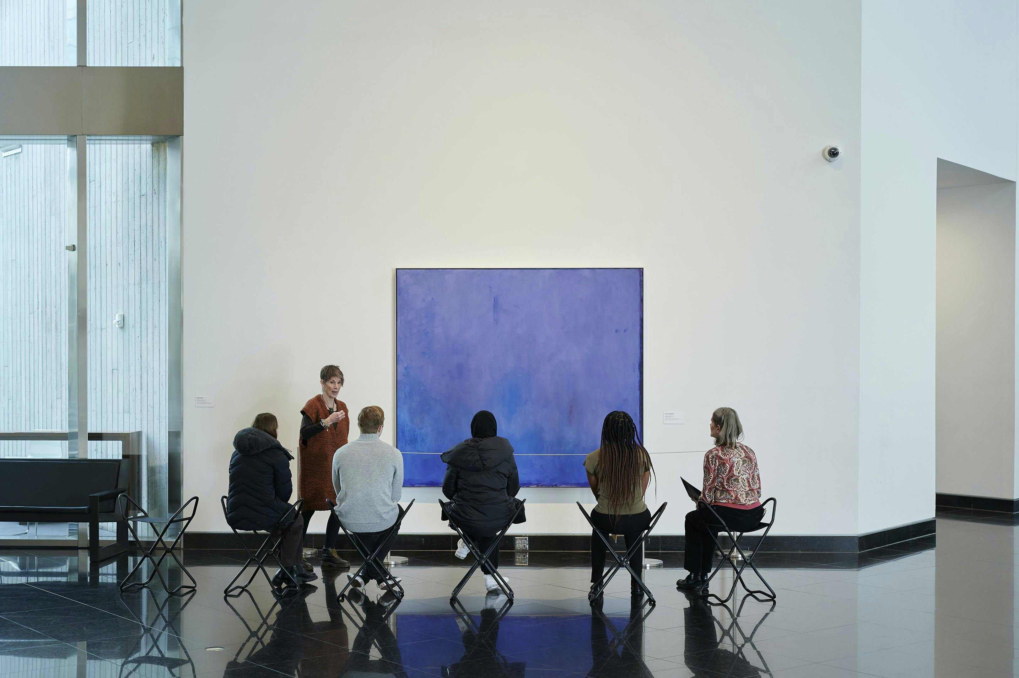 Image of a museum docent talking about a painting in front of a group of seated adults in the Kemper Museum Atrium