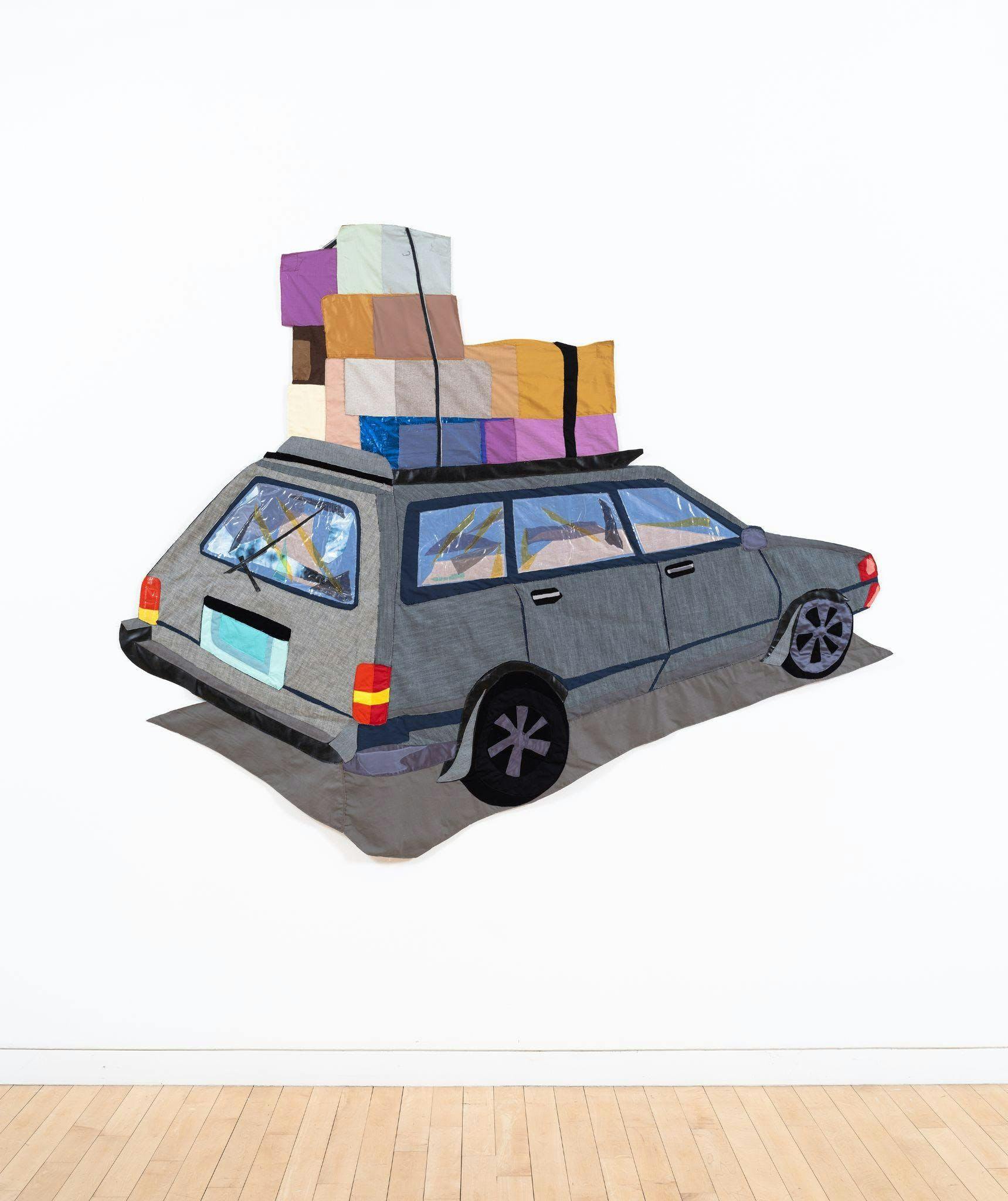Textile artwork installed in a gallery depicting a car with boxes on top.